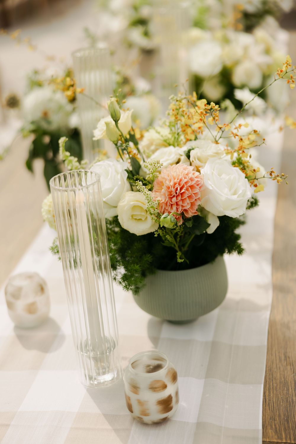 Elegant floral centerpiece with white roses and coral dahlia in sage green vase alongside crystal glassware on white tablecloth.
