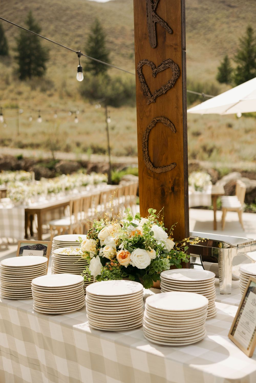 White dinner plates are stacked neatly on a buffet table with wooden pillars and floral arrangements in an outdoor setting.