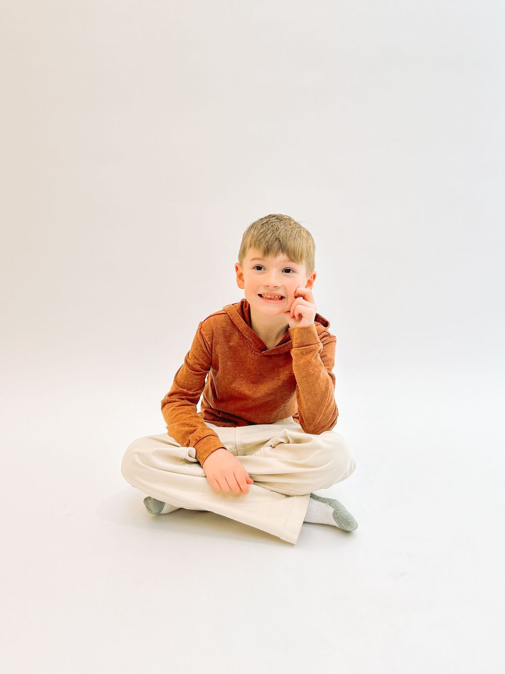 Young child in orange sweater and khaki pants sits cross-legged on white background with thoughtful expression.