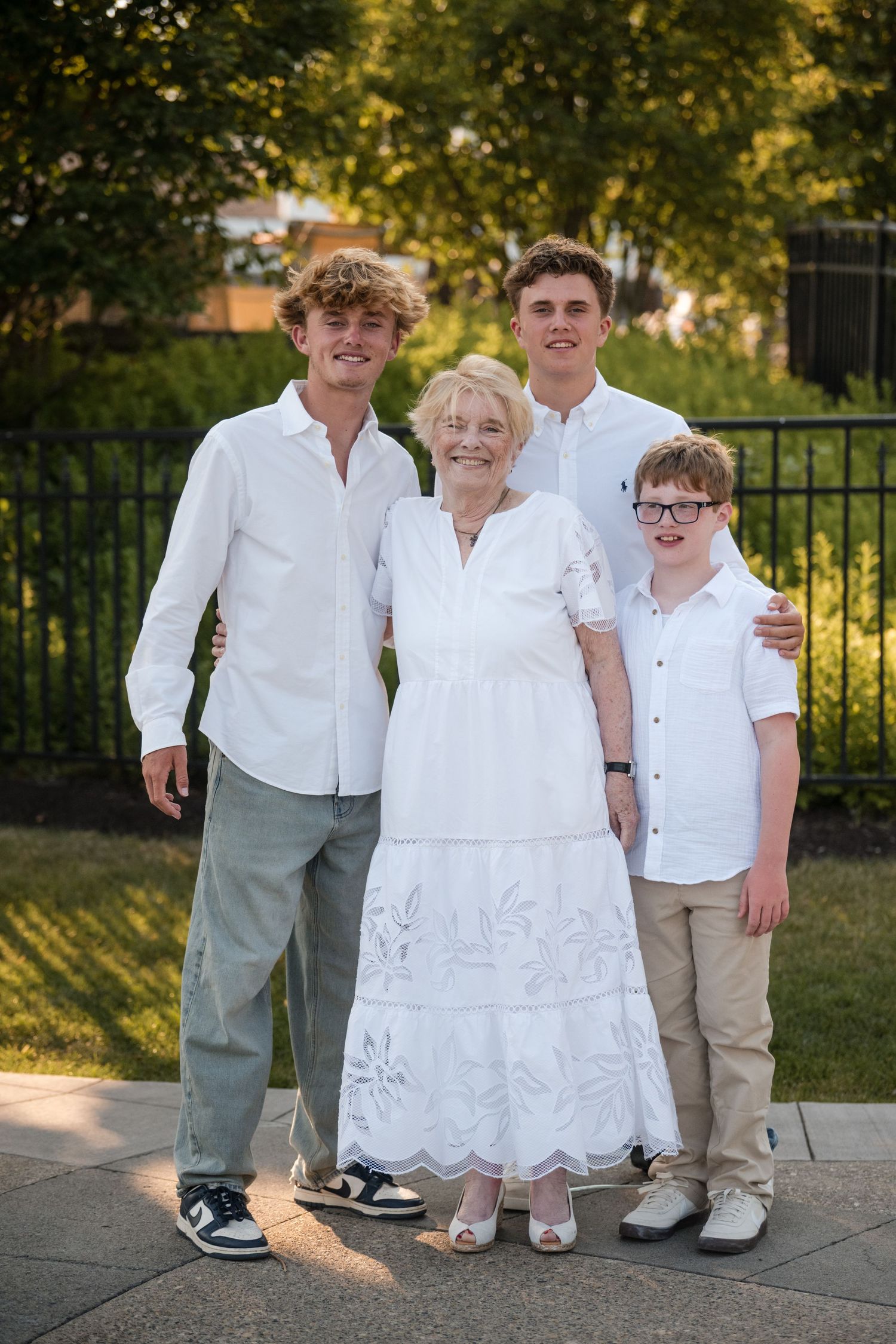 Group wearing coordinated white and khaki outfits poses together outdoors in golden sunlight near a fence.