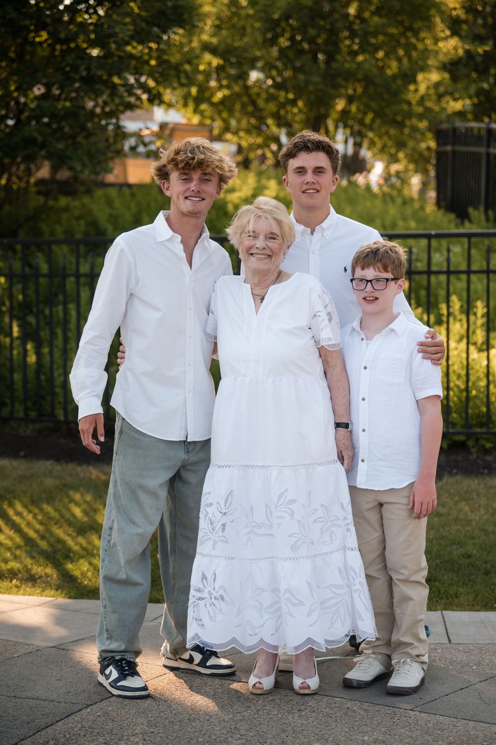 Group wearing coordinated white and khaki outfits poses together outdoors in golden sunlight near a fence.