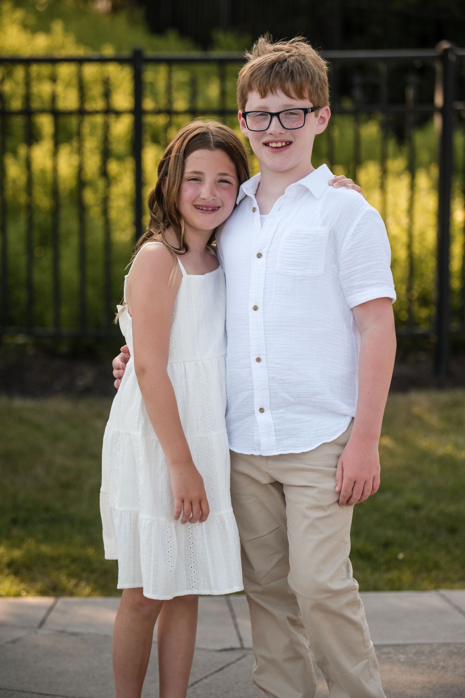 Two children in white outfits pose together outdoors on a sunny day with a fence in the background.