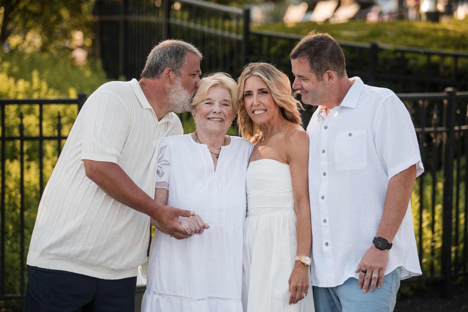 Outdoor wedding party dressed in white poses together in a garden setting at sunset.