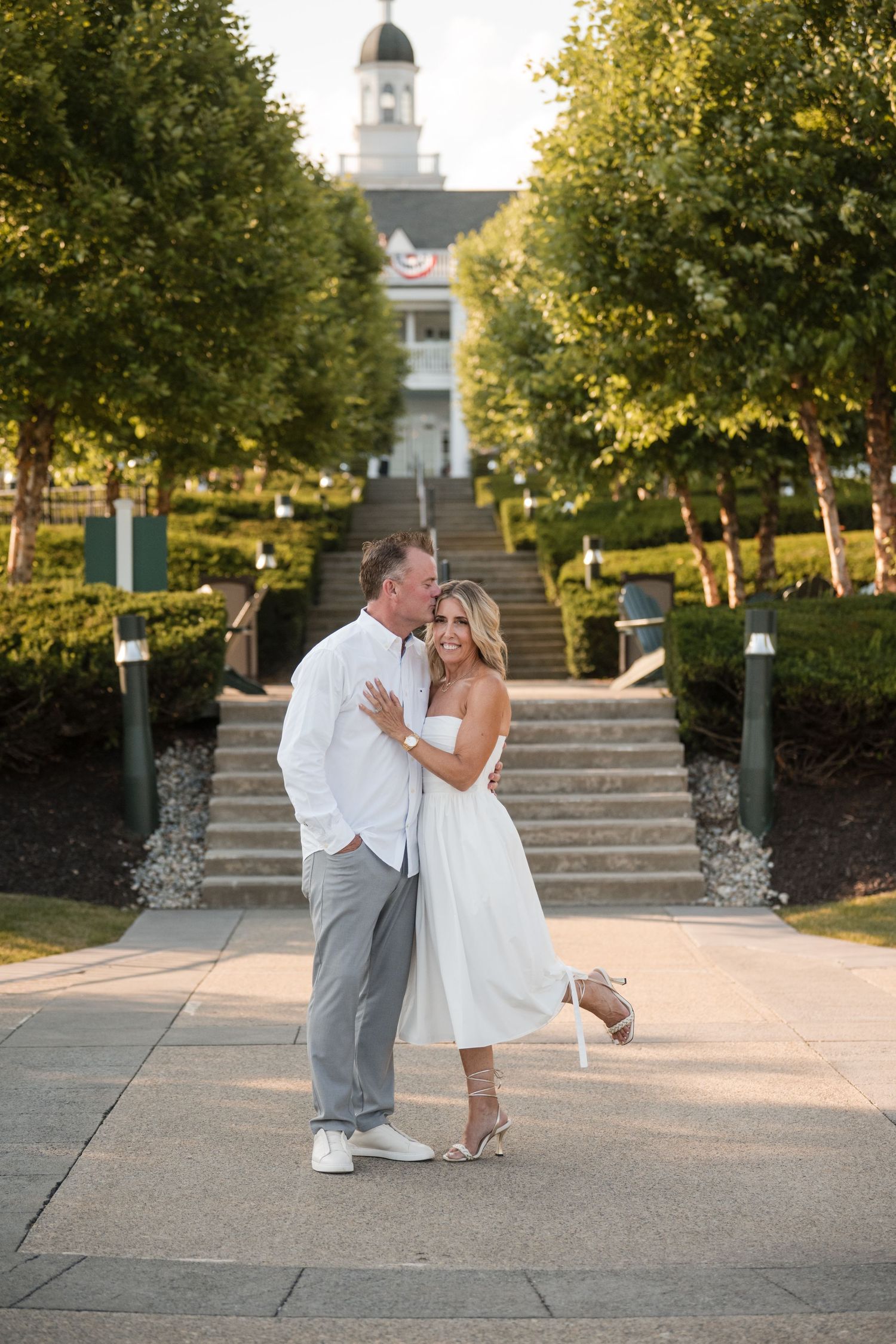 A couple shares romantic moments in white attire on stone steps with a church steeple visible in the background.