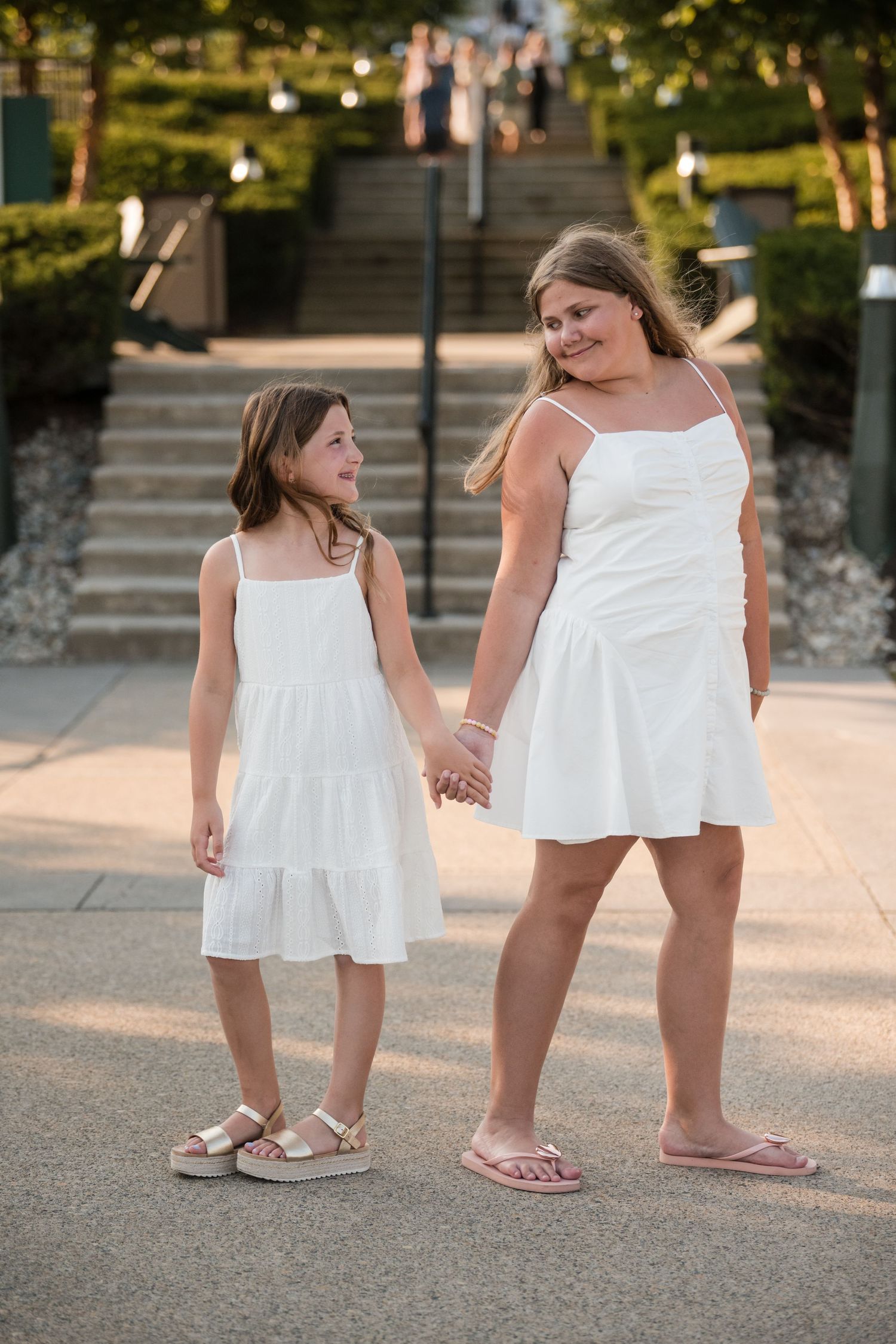 Two people in matching white summer dresses stand together holding hands on a paved path at sunset.