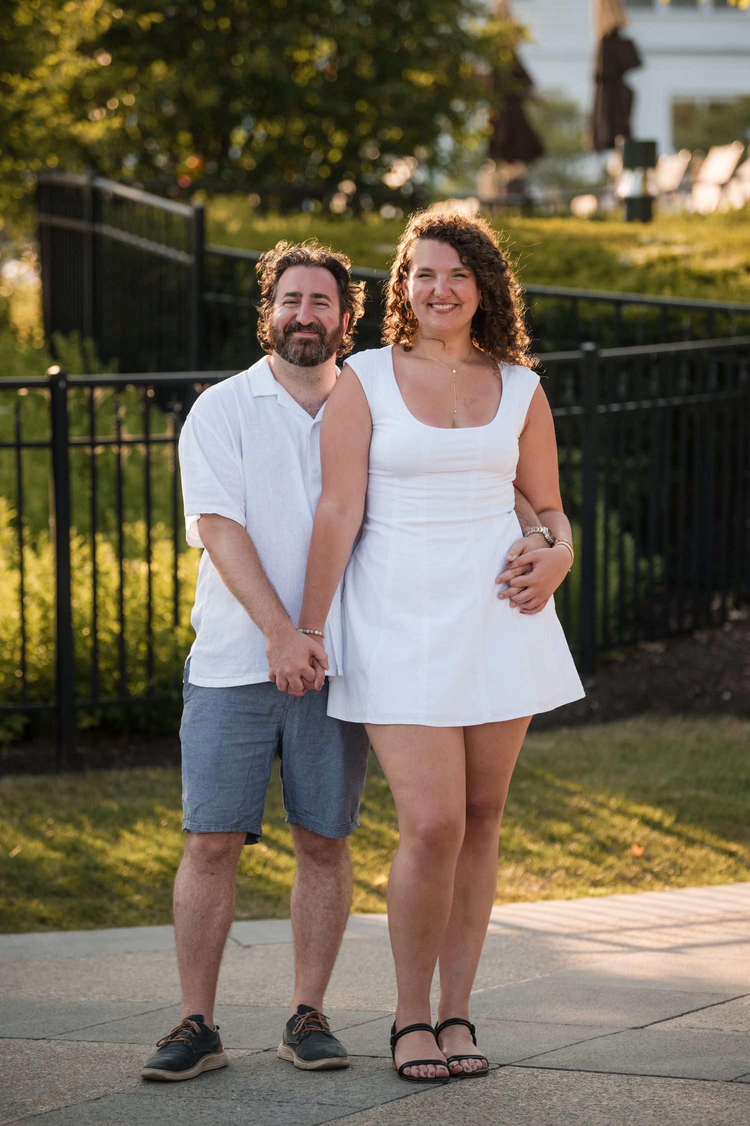 A couple in coordinating white outfits poses together on a summer evening in a park setting.