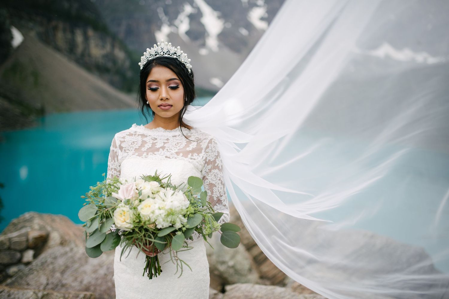 A bride in a lace gown holds a white bouquet at Moraine Lake as her veil flows dramatically in the mountain breeze.