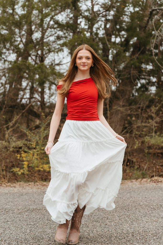 A person in a red sleeveless top and flowing white skirt poses on a country road in autumn.