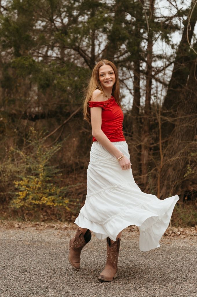 A person dances and spins in a red top and white skirt on a gravel path surrounded by autumn trees.