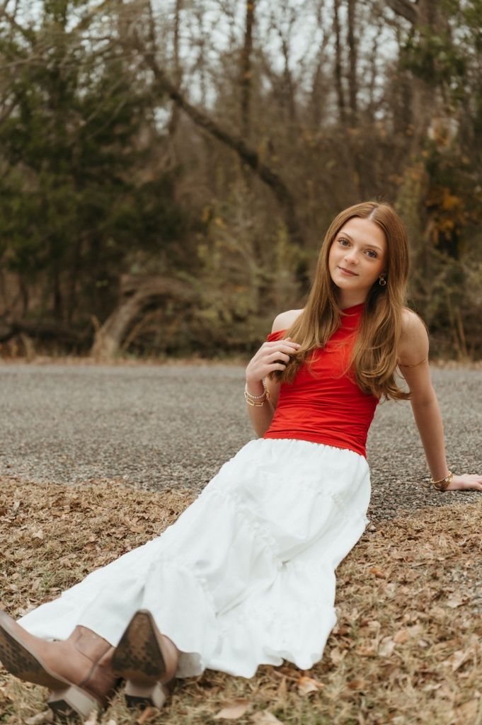 A person in a red top and white skirt poses gracefully on a bed of autumn leaves in a wooded setting.