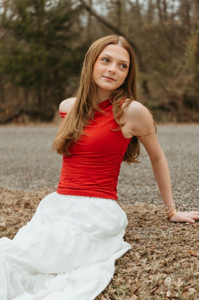 A person in a bright red sleeveless top and white flowing skirt poses playfully on fallen autumn leaves outdoors.