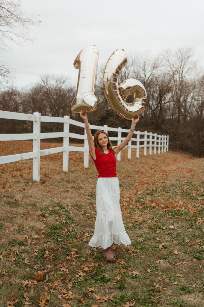 A person in a red and white dress twirls playfully near a white fence on an autumn day.