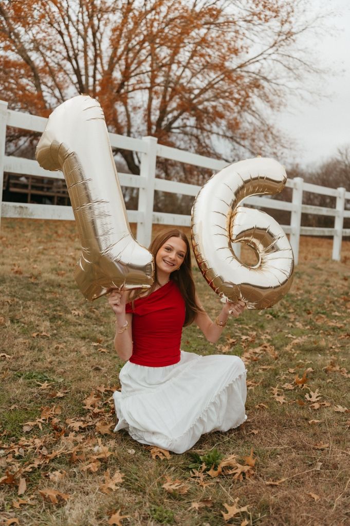 Someone in a red sweater and white skirt holds large balloon numbers while sitting on autumn ground with white fence.