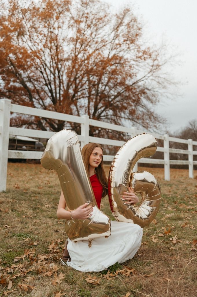 Someone in a flowing white dress lies in fallen leaves beside a rustic white fence.