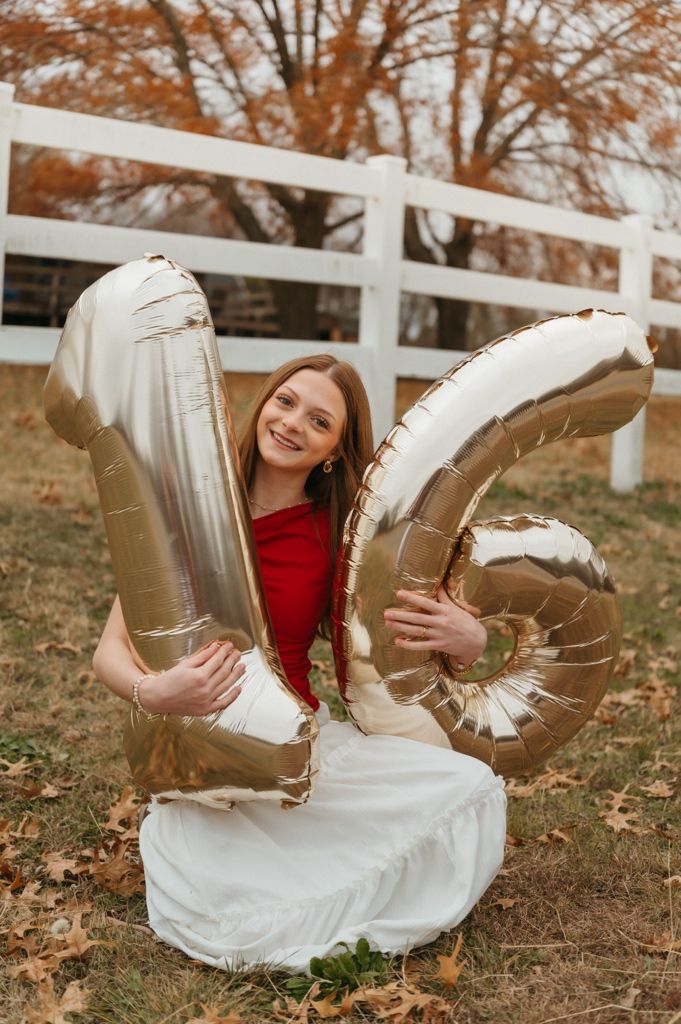 A person holds large silver number balloons while wearing a red top and white skirt outdoors.