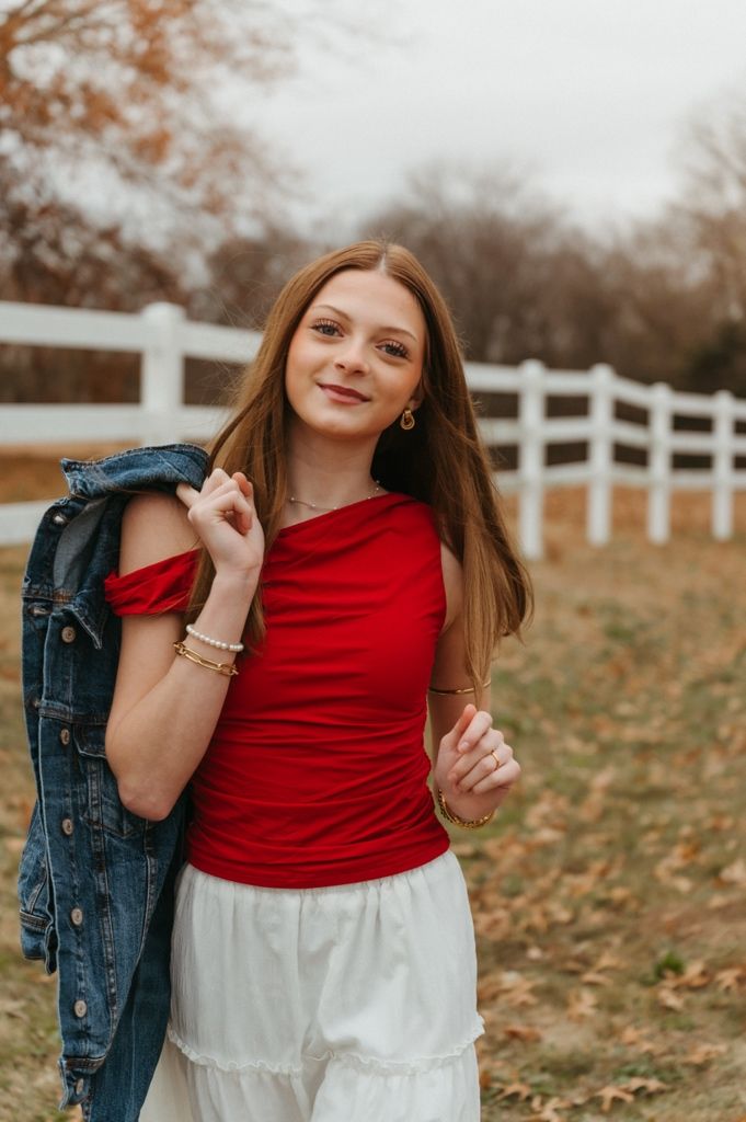 A person in a red top and white skirt poses casually with a denim jacket against a white fence in autumn.