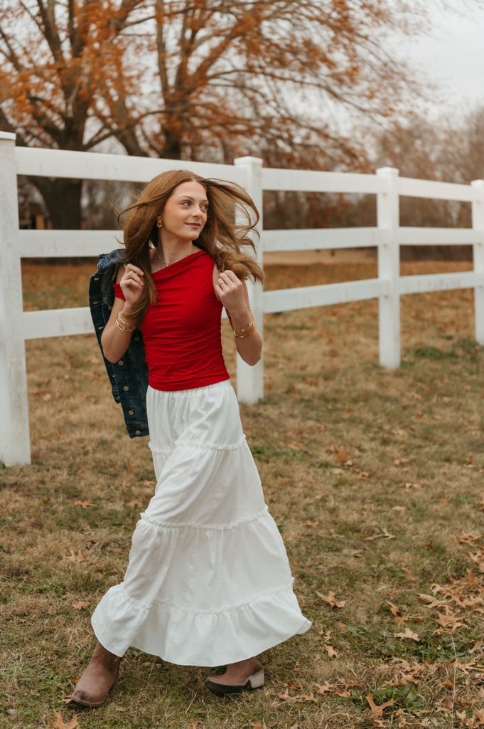 A person twirls in a flowing white maxi skirt and red top near a white fence during fall.