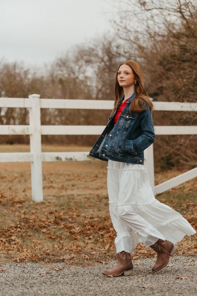 A woman in a denim jacket and flowy white skirt poses near a white fence on an autumn day.
