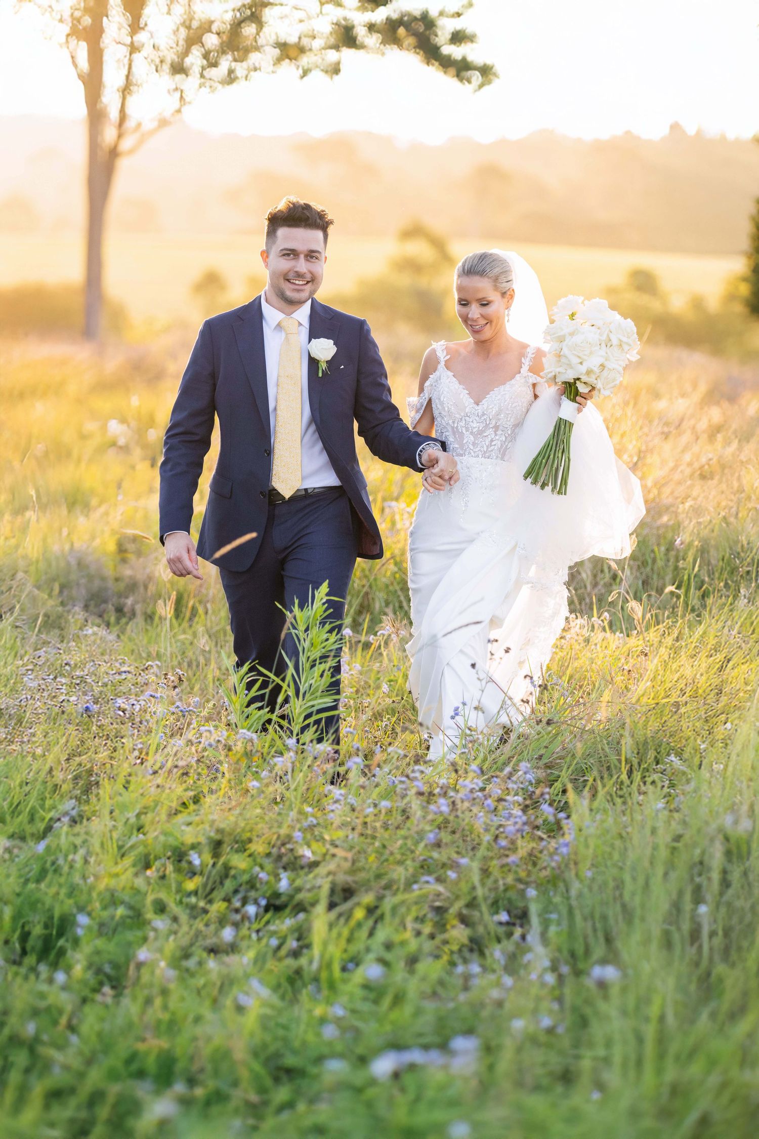 Joyful bride and groom walking hand in hand through wildflower meadow at golden sunset.