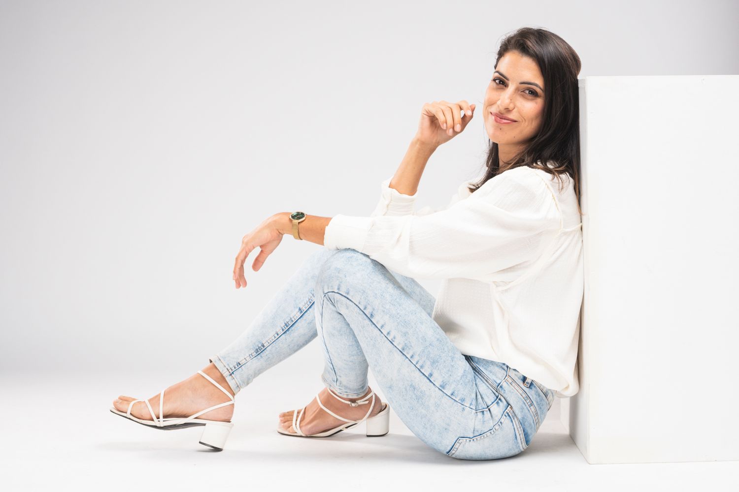 Casual fashion model wearing white shirt and distressed jeans poses sitting on floor against white background.