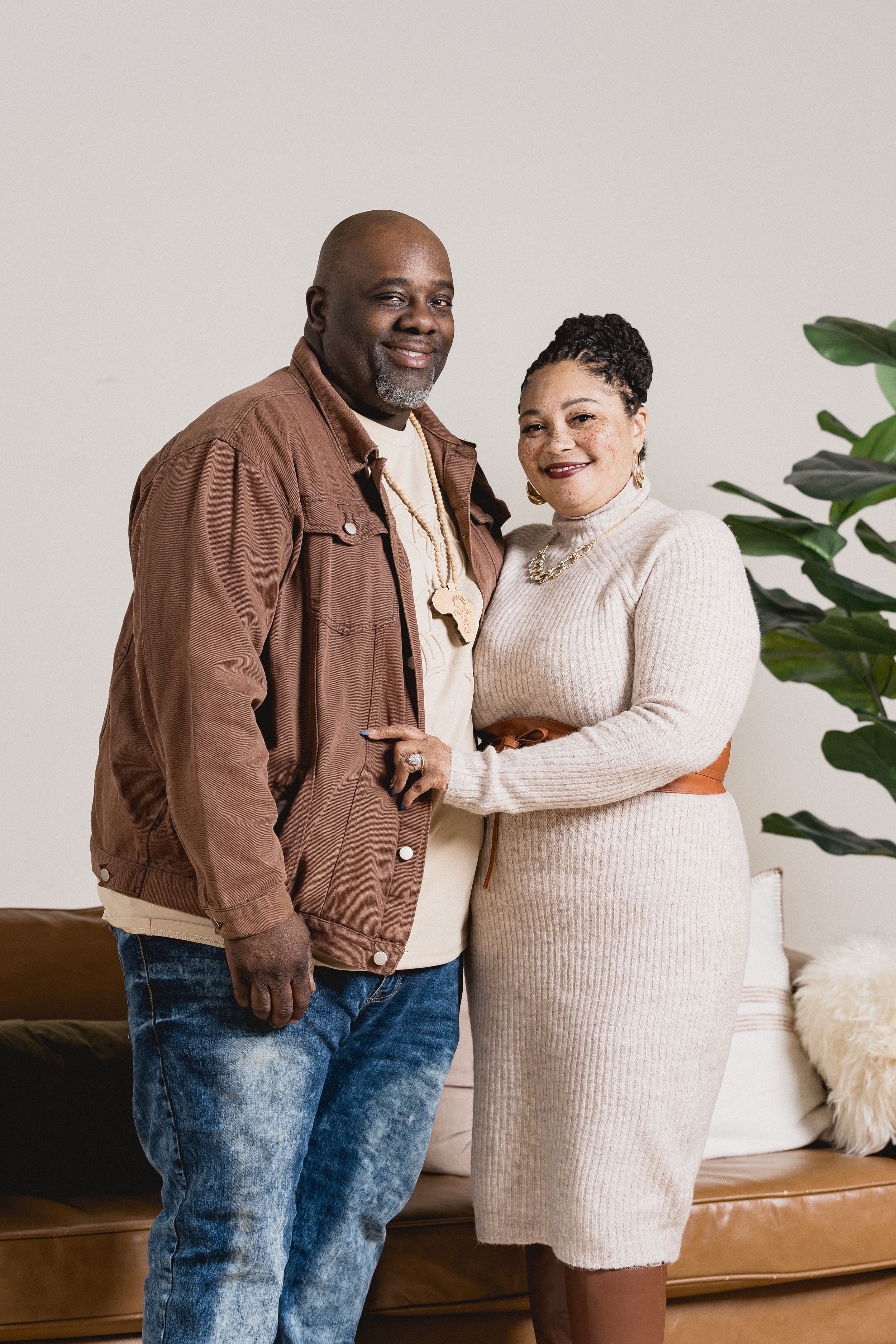 A couple in casual attire share a warm embrace against a white wall with indoor plants in the background.