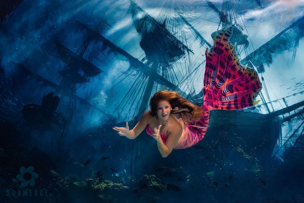 A figure in a flowing red dress floats underwater near a sunken ship in a mysterious blue underwater scene.