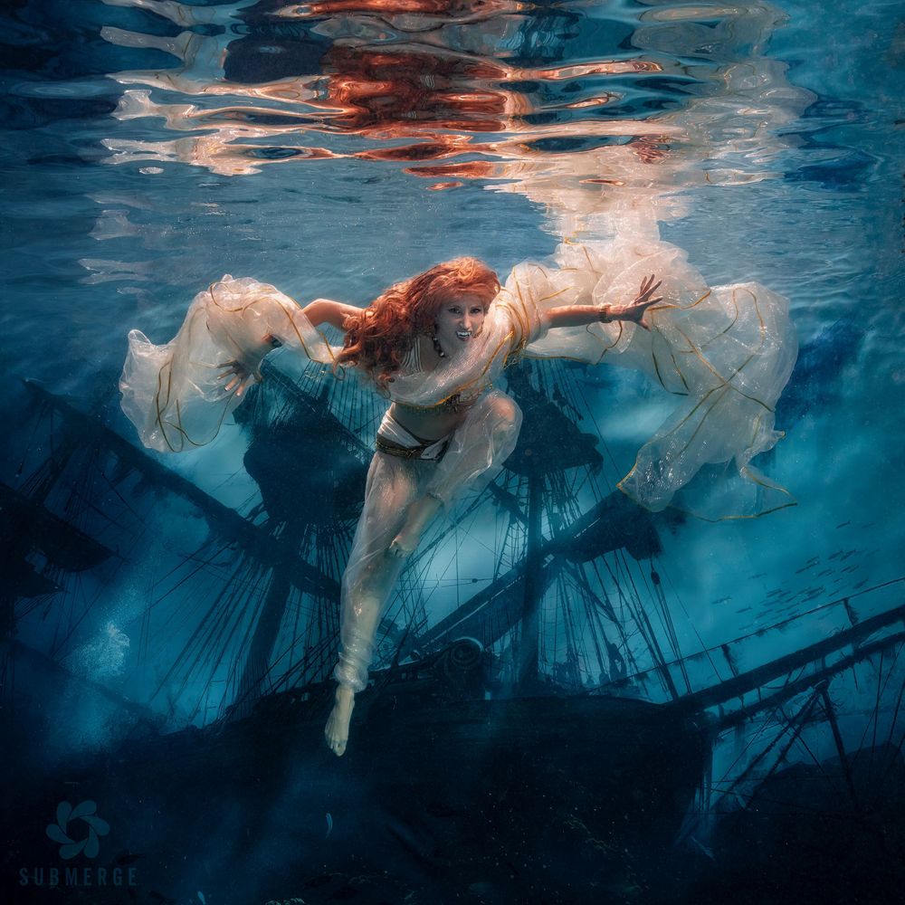 Underwater view of a dancer in a flowing blue dress creating ethereal movements beneath the surface of a pool.