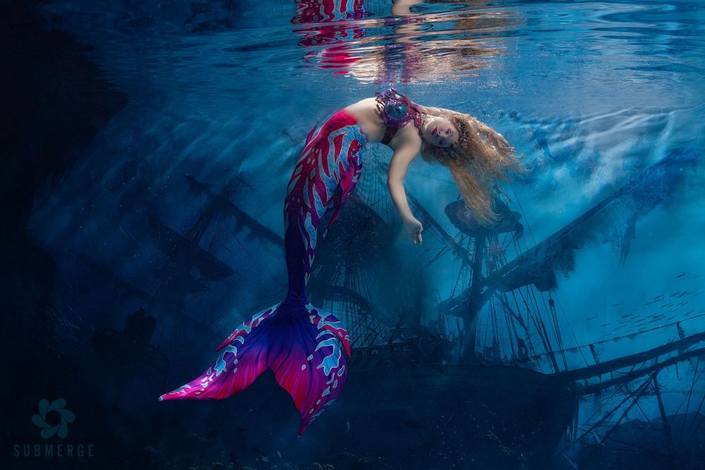 A figure in a flowing pink dress moves gracefully underwater against a deep blue background creating an ethereal scene.