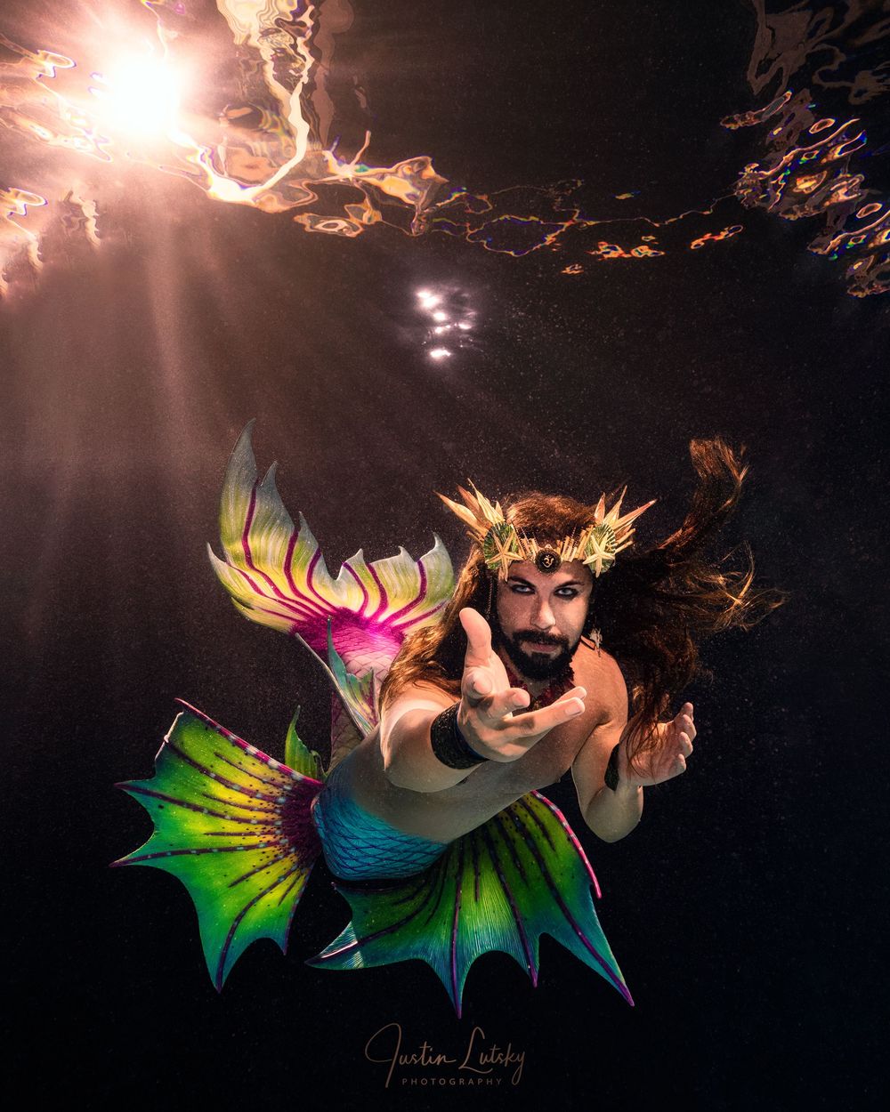 Underwater fantasy figure wearing colorful mermaid tail and crown poses dramatically with light beams in dark water.