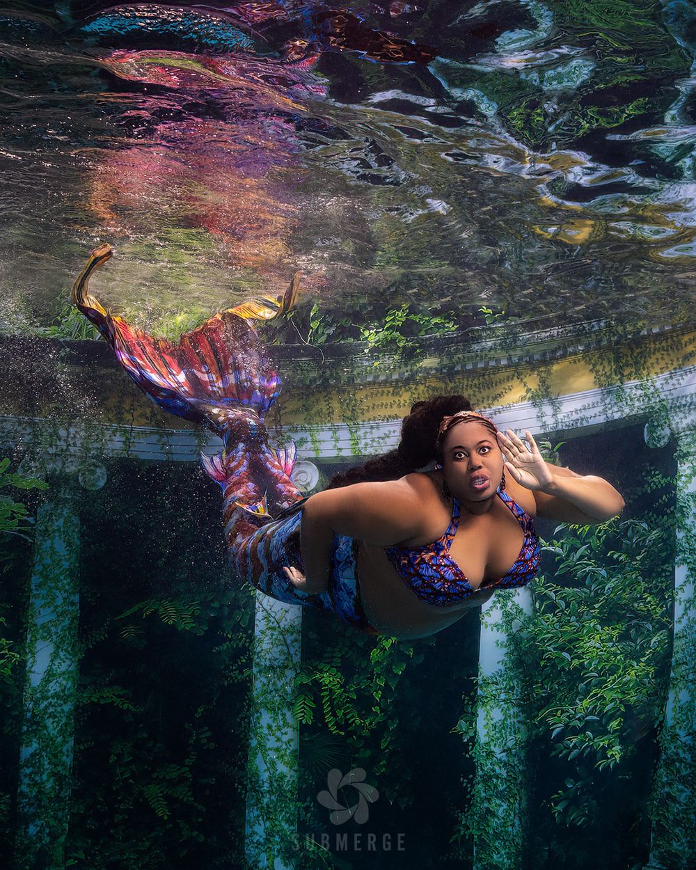Underwater photograph showing a figure in a purple swimsuit gracefully floating beneath the surface with colorful reflections above.