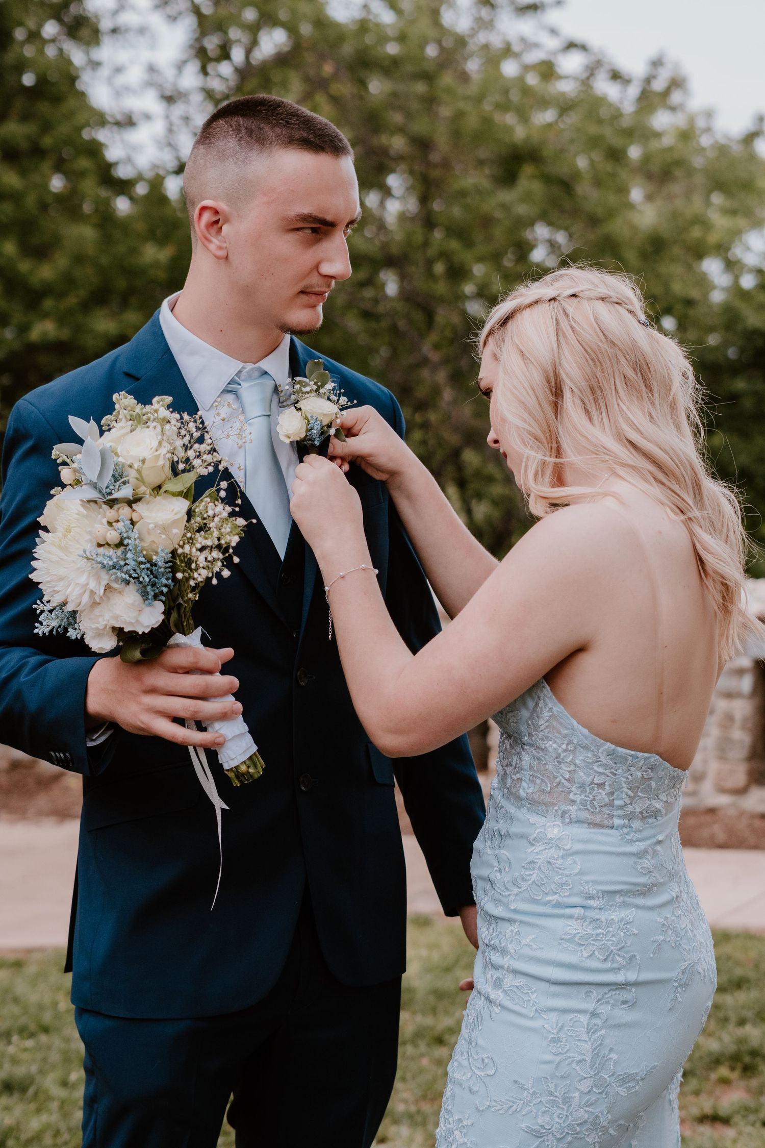 A couple in wedding attire share an intimate moment outdoors while the bride adjusts the groom's boutonniere.