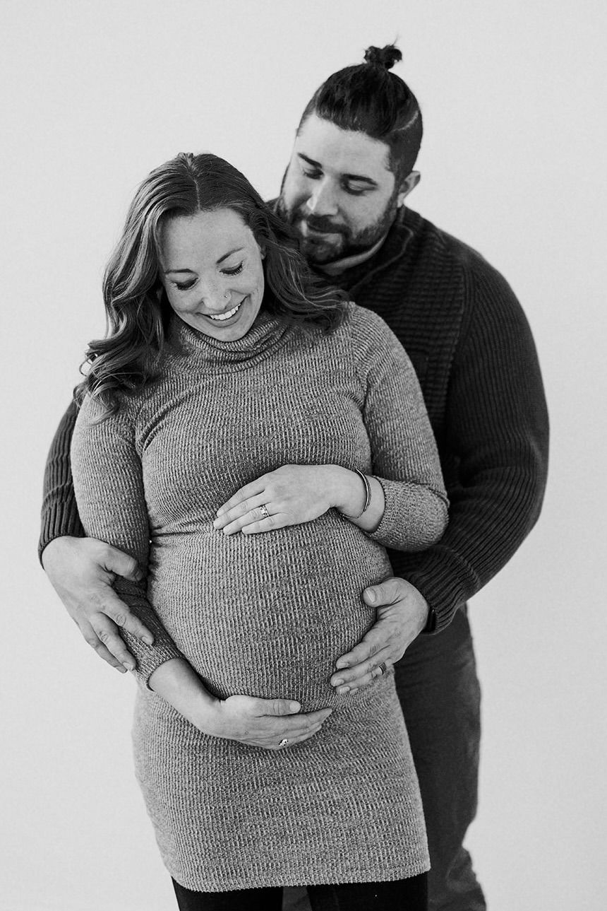 Maternity photoshoot featuring couple in grey sweater embracing baby bump against white background.