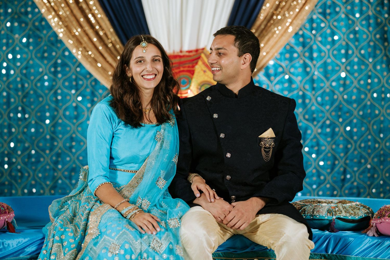 A couple in traditional Indian attire shares a joyful moment against a sparkly turquoise backdrop during a celebration.