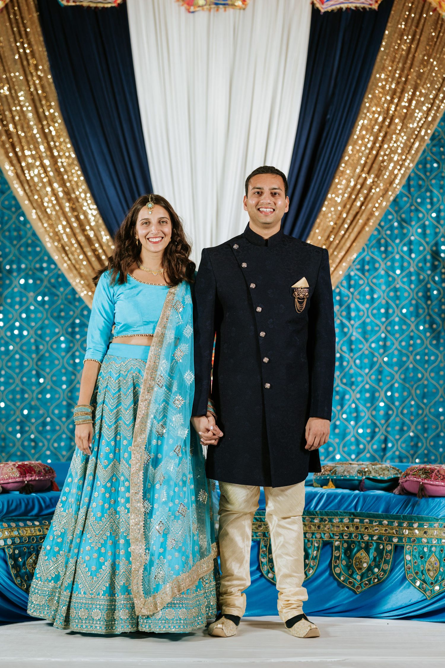 A couple in traditional Indian wedding attire poses against a backdrop with gold sequins and blue draping.