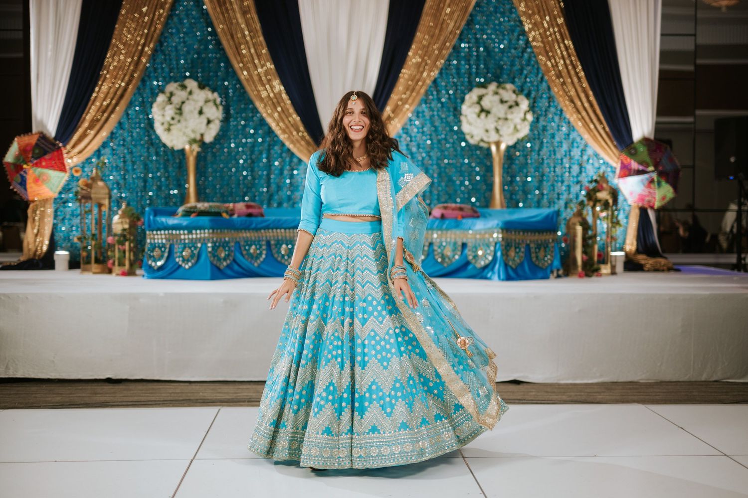 A turquoise and gold embroidered lehenga dress twirls in front of an ornate backdrop with white floral arrangements.