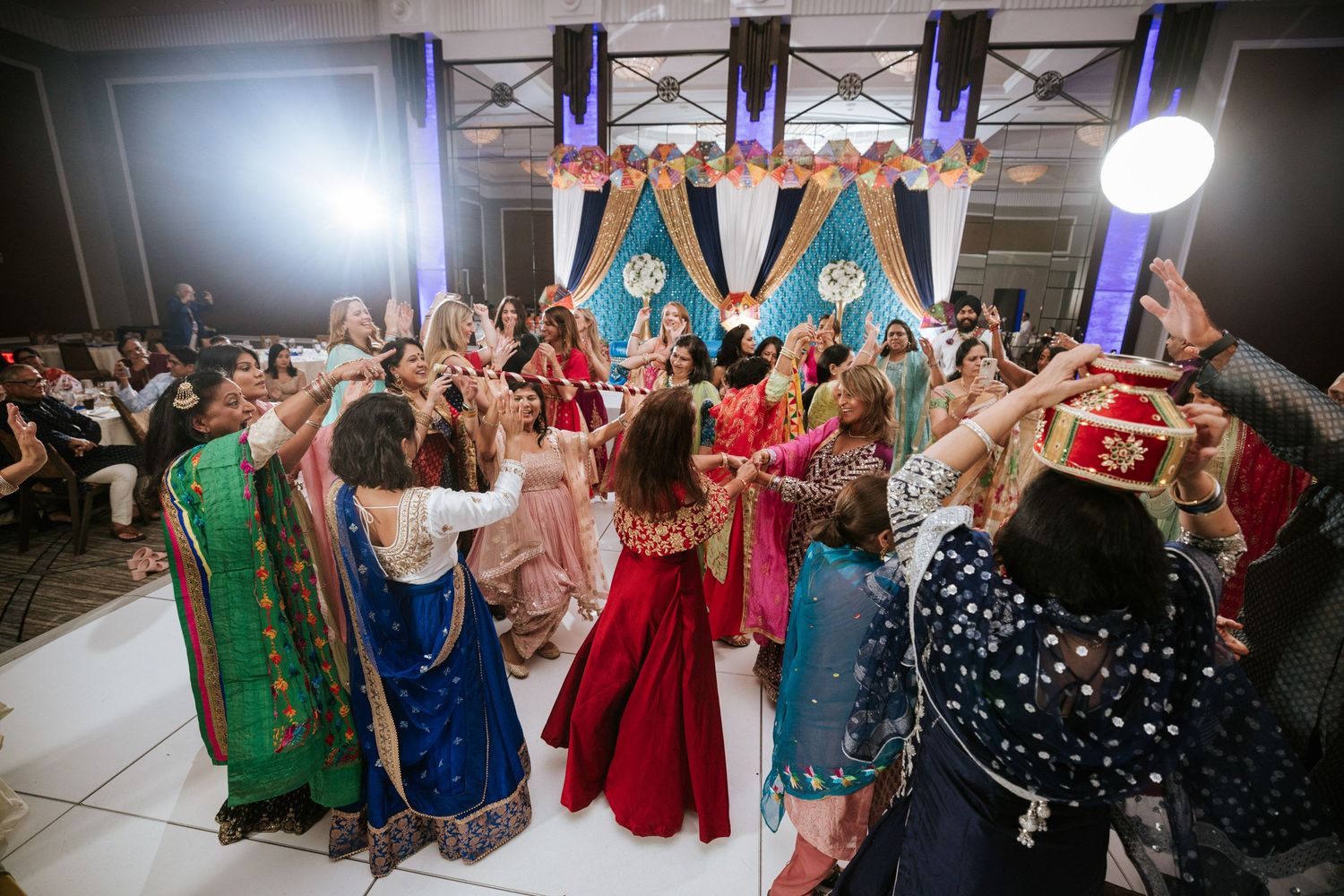 Wedding reception dance floor filled with guests in colorful traditional outfits celebrating with raised arms.