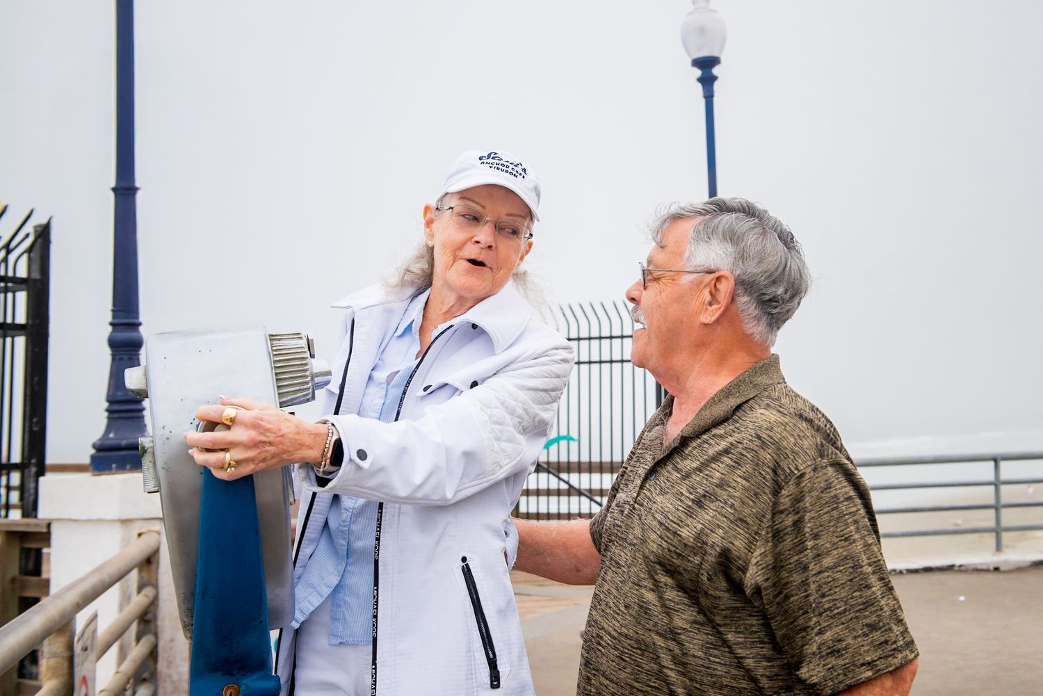 Two people enjoying conversation while standing on a pier near the ocean with a lamp post visible in the background.