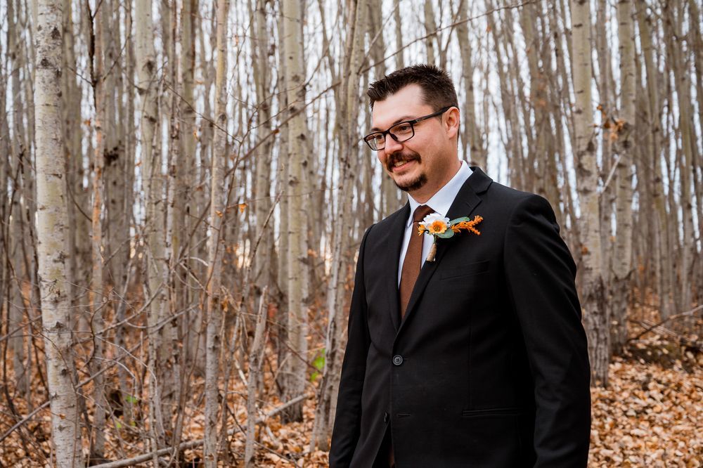 A couple in formal wedding attire stands in a forest with bare trees and fallen autumn leaves. Parkland county alberta