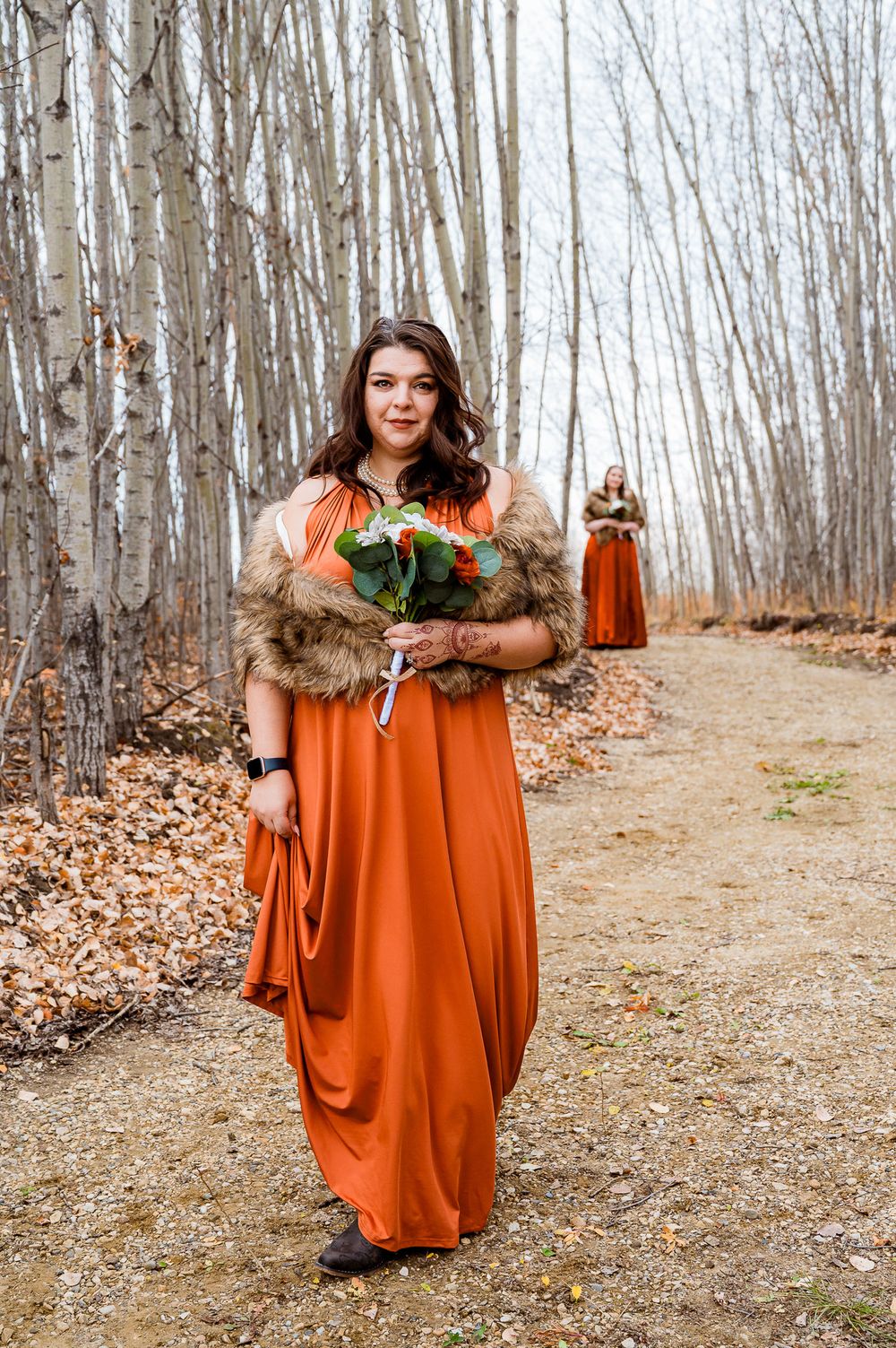 A person in a flowing orange dress poses in a serene birch forest setting with fallen autumn leaves on the ground. Bridesmaid edmonton