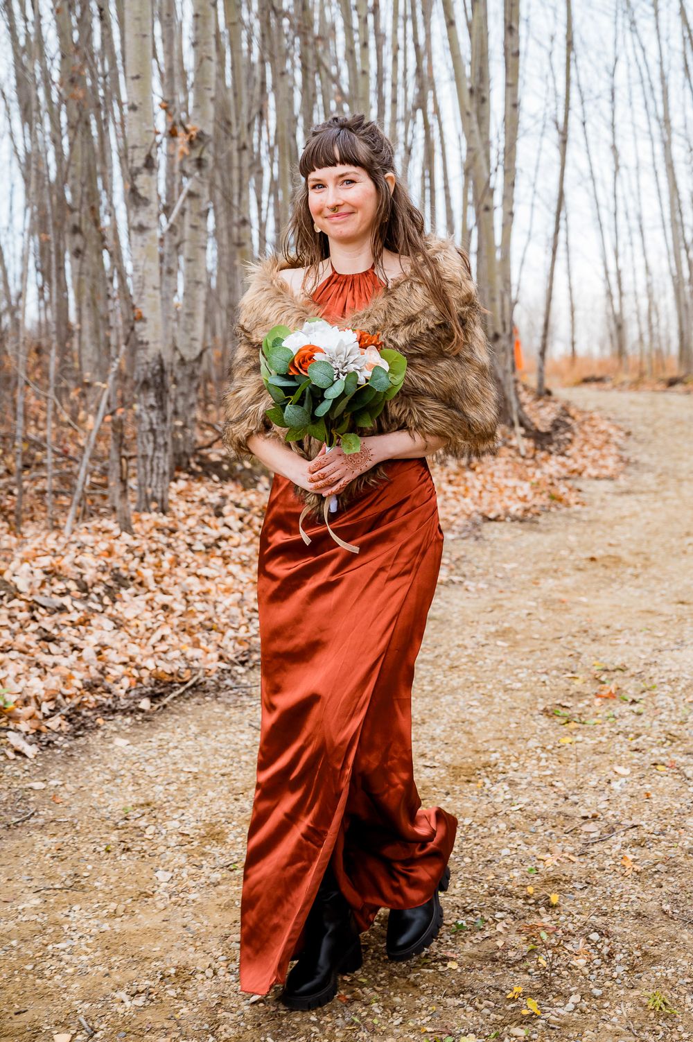 A person in a flowing rust-colored dress twirls through autumn woods holding flowers on a leaf-covered forest path. edmonton bridesmaid
