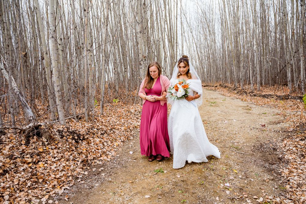 Two people in formal dresses walk along a forest path covered in autumn leaves with bare trees in the background. village vows seba beach
