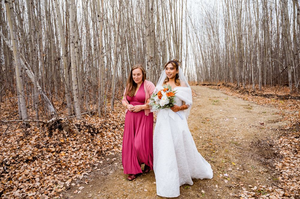 A group walks along a wooded path in autumn with burgundy and white dresses amid fallen leaves. edmonton wedding ceremony, seba beach