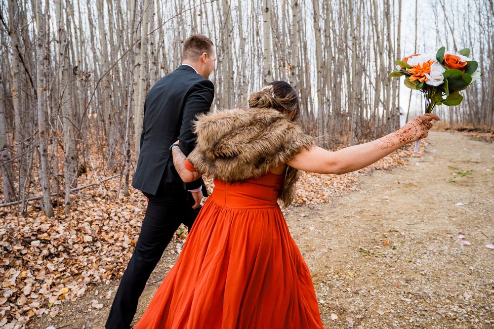 A couple in formal attire walks through a forest path during autumn with orange dress and black suit. Seba beach wedding ceremony