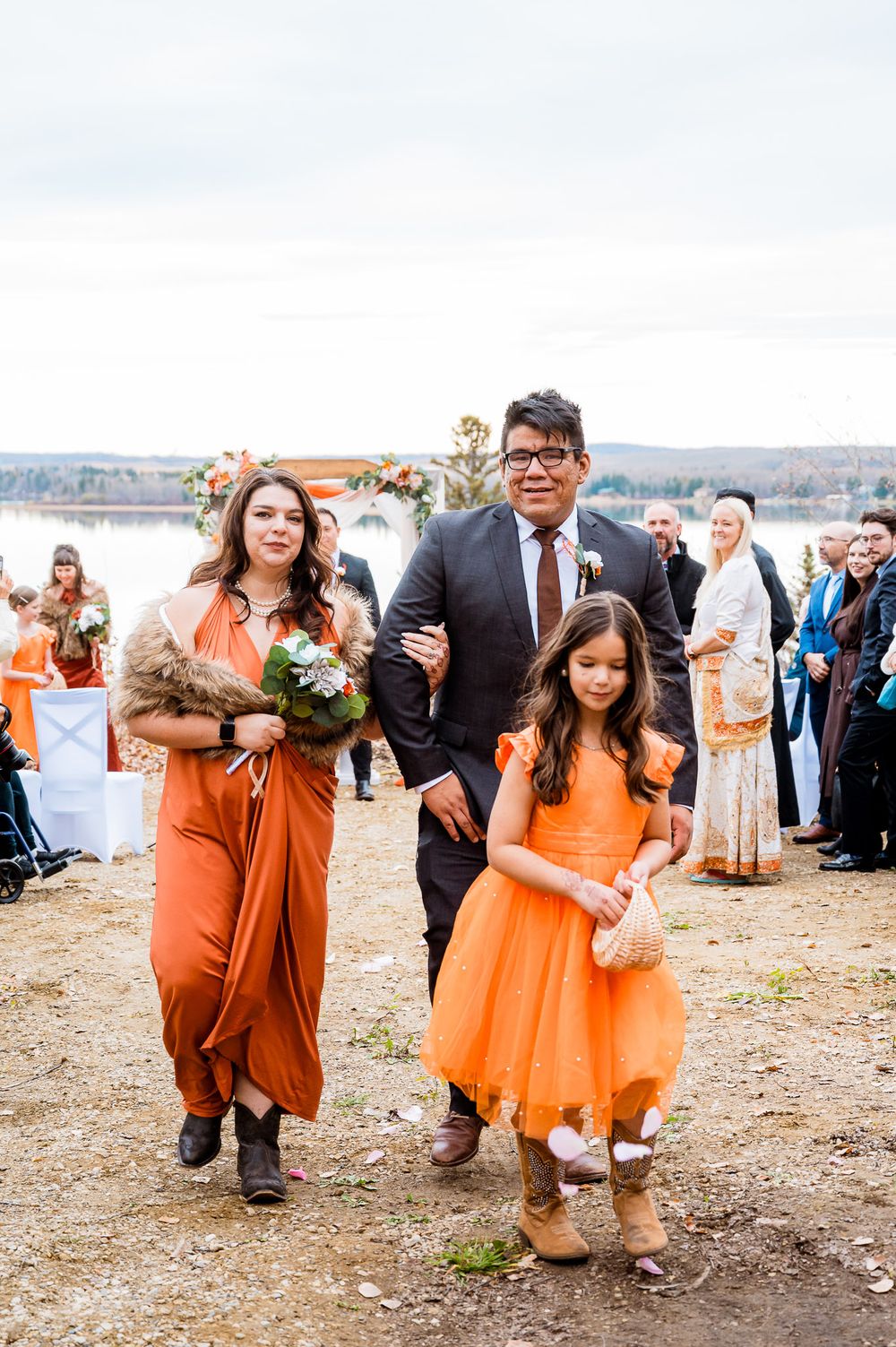 Wedding party members in vibrant orange dresses and suits walk together outdoors during a fall celebration. Seba Beach wedding videographer