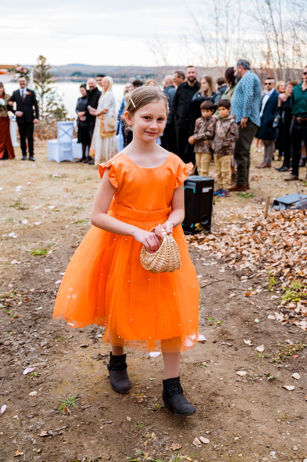A young attendant in a bright orange tulle dress carries a basket at an autumn outdoor wedding celebration. forest lodge village vows