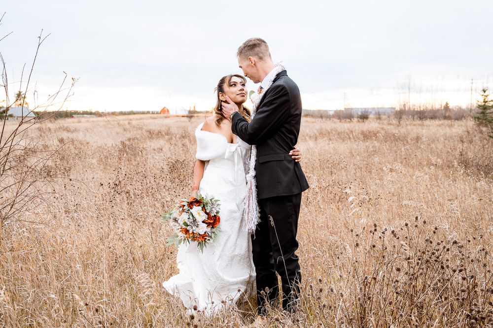 A couple in wedding attire embraces in a rustic field of tall grass while holding a colorful autumn bouquet. village vows seba beach wedding