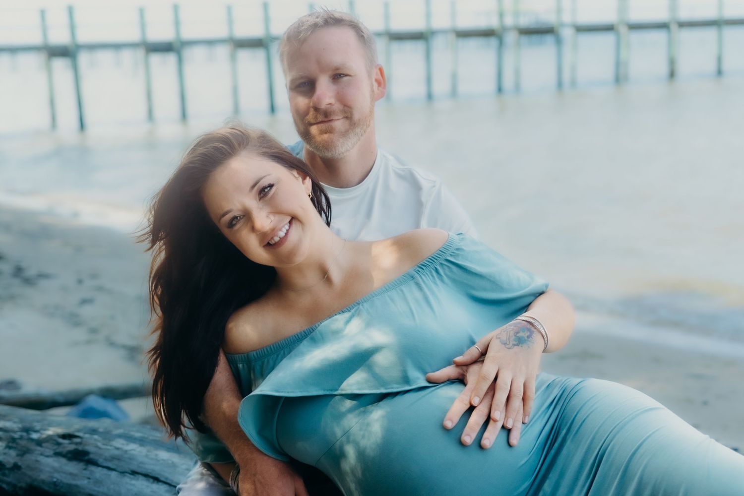 Couple in casual attire posing together on beach with pier in background.