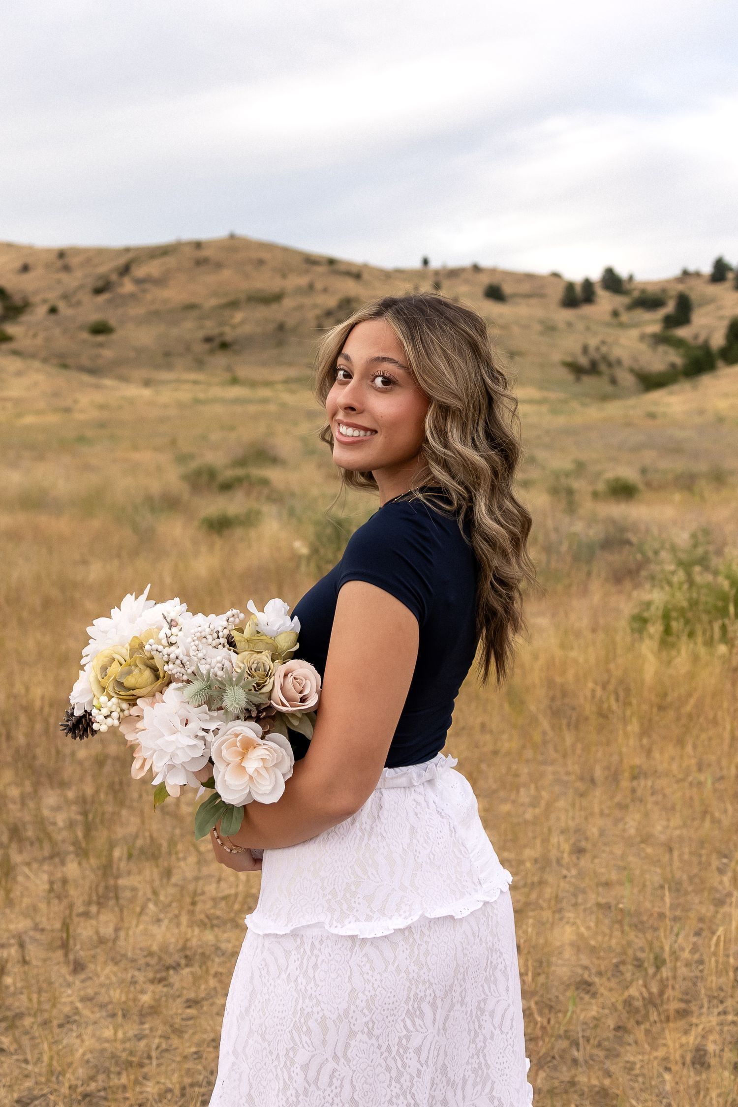 A person in a white dress holds a bouquet of white flowers while standing in a golden grassy field on a sunny day.