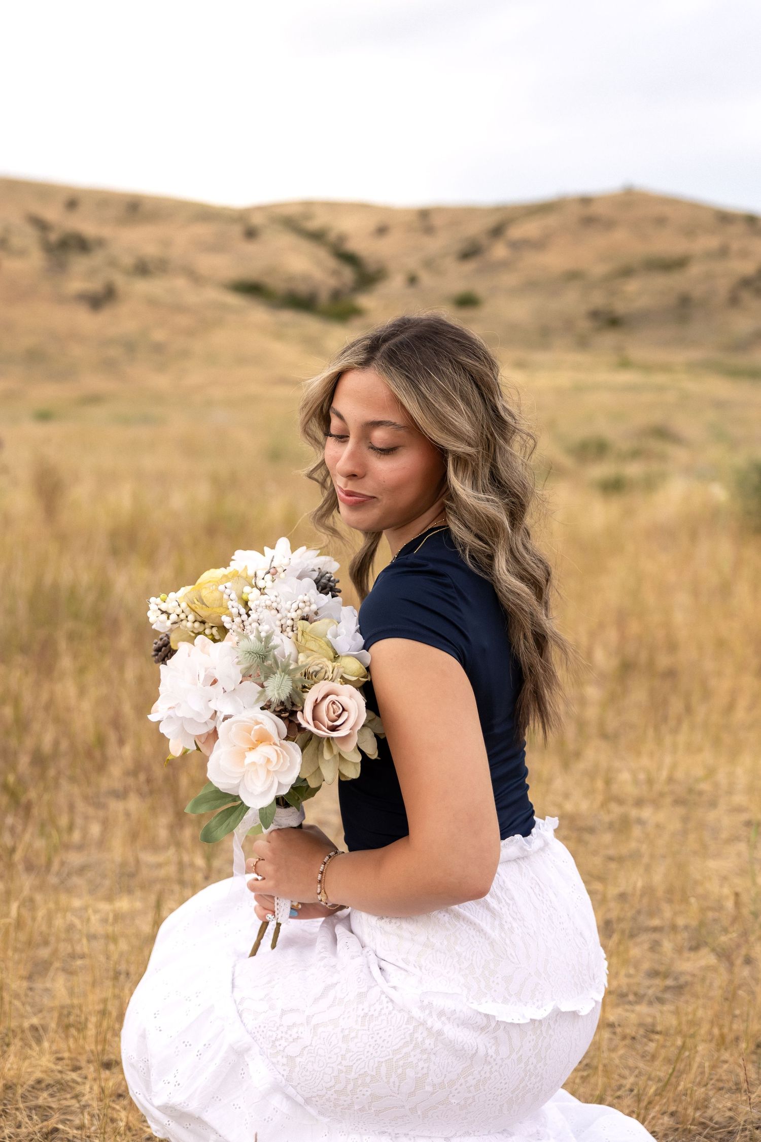 A woman in a flowing white skirt poses with a delicate floral bouquet against rolling hills at sunset.