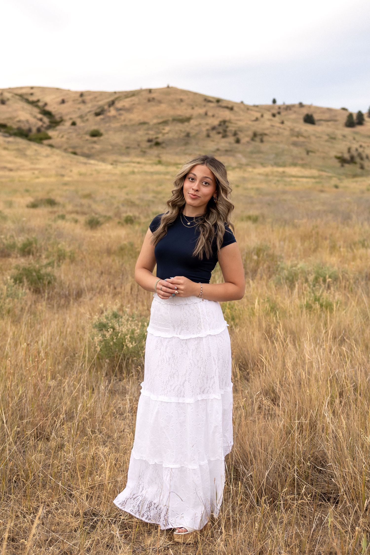 A person in a black top and white tiered skirt poses gracefully in a golden autumn field against rolling hills.